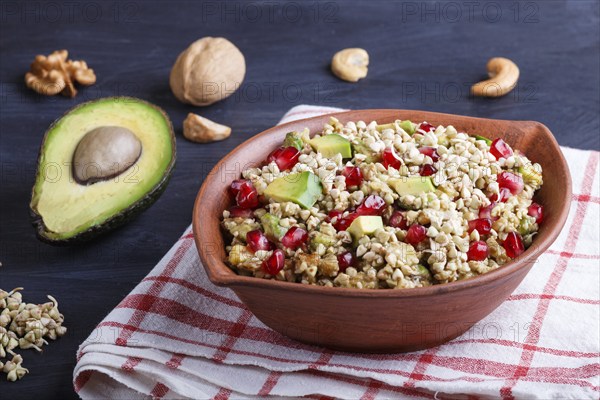 Salad of germinated buckwheat, avocado, walnut and pomegranate seeds in clay plate on black wooden background. Side view