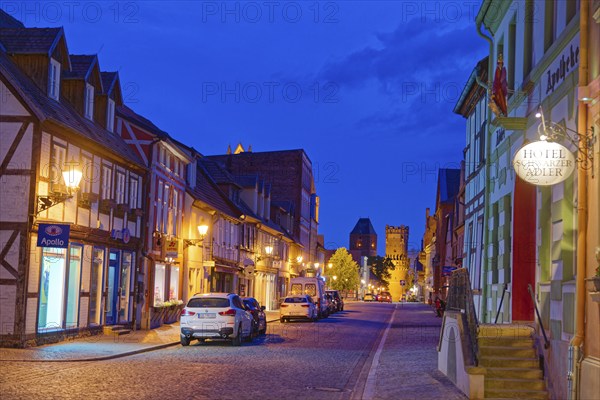 The Lange Straße with half-timbered houses and other old buildings, covered with cobblestones, illuminated in the evening. The Neustädter Tor in the background. Old town centre of Tangermünde, Hanseatic town in the Altmark. Saxony-Anhalt, Germany, Europe