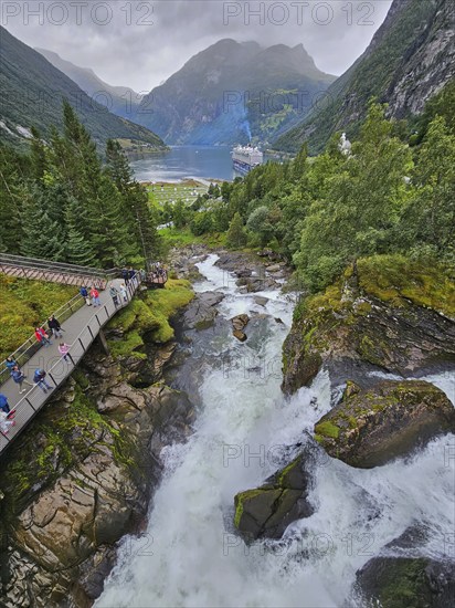 An impressive landscape with a raging waterfall, surrounded by green mountains and forests, with a cruise ship, Mein Schiff 6, on a fjord hiking trail with walkway and tourists, Geiranger, Geiranger Fjord, Stranda, Romsdal, Norway, Europe