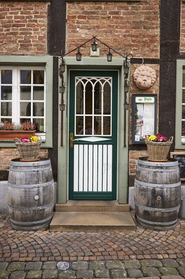 Entrance area of a restaurant in a historic half-timbered brick house with entrance door, white windows, old wooden barrels and flower baskets for decoration in Warendorf, Warendorf district, North Rhine-Westphalia, Germany, Europe