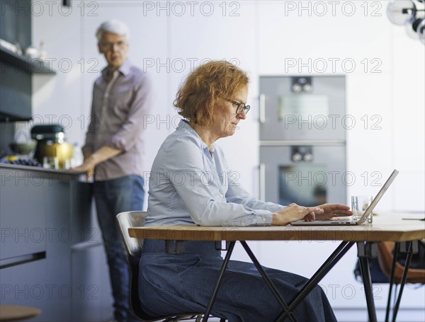 Symbolic photo on the subject of division of labour for couples in the household. A woman sits at a laptop in a kitchen while a man washes dishes in the background. Berlin, 13.08.2024