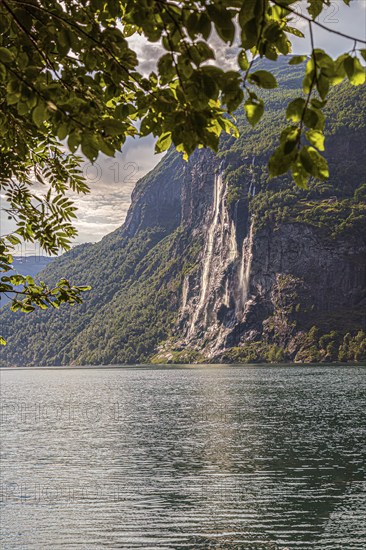 The majestic Seven Sisters waterfall flows down a forested mountain into the tranquil Gairanger Fjord under a partly cloudy sky in Norway Europe