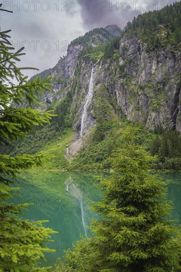 Picturesque waterfall that cascades majestically along a rocky cliff into an emerald green lake, surrounded by dense forest, Stilluptal, Zillertal. Austria