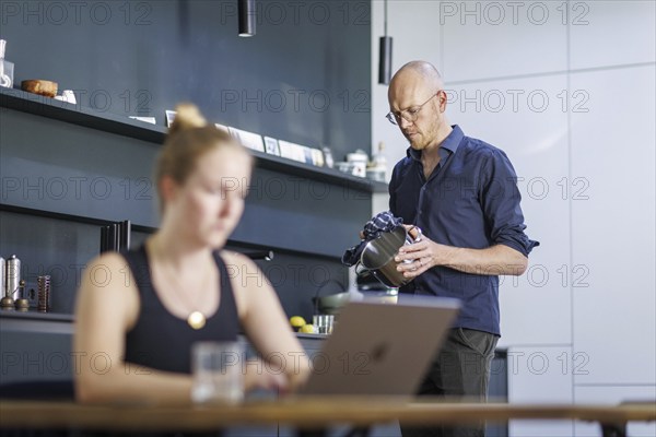 Symbolic photo on the subject of division of labour for couples in the household. A woman sits at a laptop in a kitchen while a man washes dishes in the background. Berlin, 13.08.2024