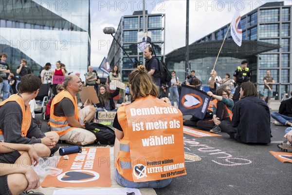 Last Generation activist with poster Democracy is more than voting! for the upcoming European elections at the occupation of Rahel-Deer-Straße in front of Berlin Central Station, Berlin, 25 May 2024