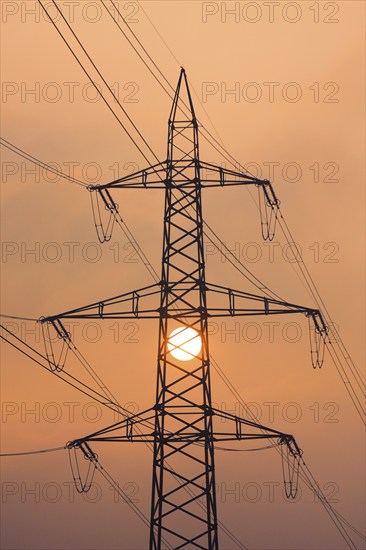 Setting sun behind silhouette of high-voltage power line pylons, shrouded in orange light amidst fog, Canton Zurich, Switzerland, Europe