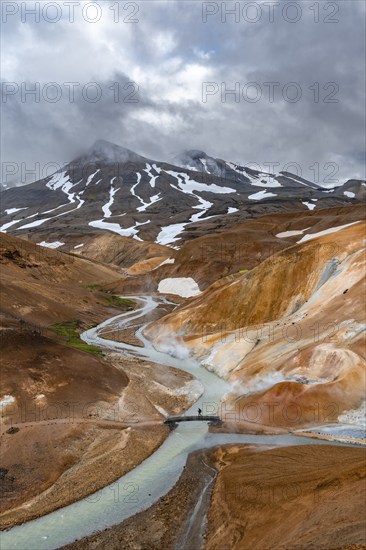 Tourist on bridge over steaming stream between colourful rhyolite mountains with snowfields, Hveradalir geothermal area, Kerlingarfjöll, Icelandic highlands, Iceland, Europe