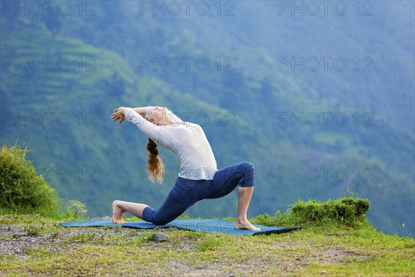 Yoga outdoors, sporty fit woman practices Hatha yoga asana Anjaneyasana, low crescent lunge pose posture outdoors in Himalayas mountains