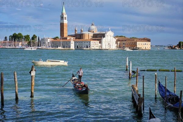 VENICE, ITALY, JUNE 27, 2018: Gondolier with client tourists in gondola in lagoon of Venice by Saint Mark (San Marco) square with San Giorgio di Maggiore church in Venice from Ponte della Paglia