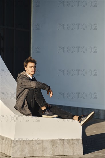 Young man in grey coat, black jumper, white shirt and black jeans sits on the street