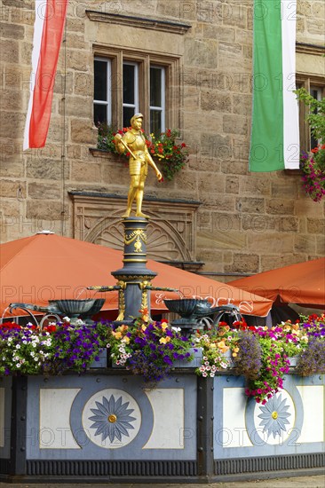 Margrave George Fountain, polygonal iron basin, gilded knight figure stands on richly decorated pillar with four shell bowls, erected by Margrave George in 1515, flower boxes, parasols at the back of town hall, Ansbach, Middle Franconia, Franconia, Bavaria, Germany, Europe