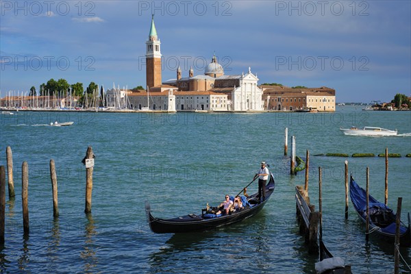 VENICE, ITALY, JUNE 27, 2018: Gondolier with tourists in gondola in lagoon of Venice by Saint Mark (San Marco) square with San Giorgio di Maggiore church in background in Venice, Italy, Europe