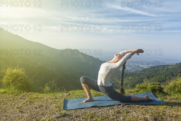 Sporty fit woman practices yoga Anjaneyasana, low crescent lunge pose outdoors in mountains in morning