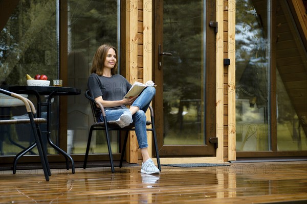 Happy woman sits in wicker chair at wooden terrace and reading the book