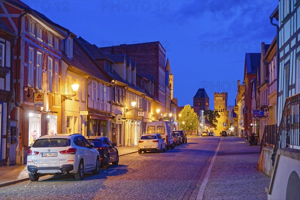 The Lange Straße with half-timbered houses and other old buildings, covered with cobblestones, illuminated in the evening. The Neustädter Tor in the background. Old town centre of Tangermünde, Hanseatic town in the Altmark. Saxony-Anhalt, Germany, Europe