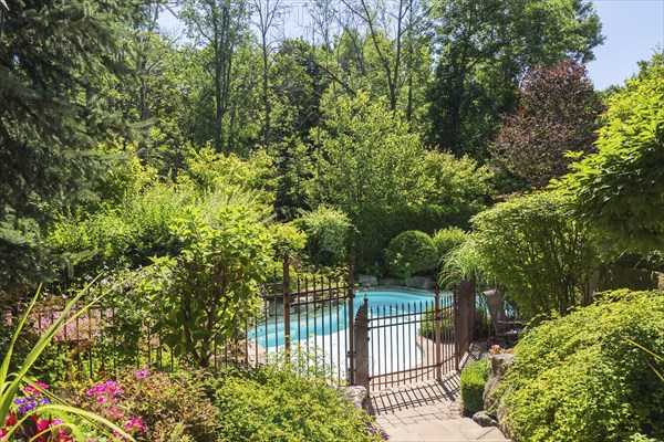 In-ground swimming pool surrounded by copper coloured wrought iron metal security fence and various perennial plants, shrubs and trees in backyard of luxurious home, Quebec, Canada, North America