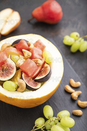 Vegetarian fruit salad of watermelon, grapes, figs, pear, orange, cashew on slate board on a black concrete background. Side view, close up, selective focus