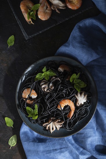 Black cuttlefish ink pasta with shrimps or prawns and small octopuses on black concrete background and blue textile. Low key, hard light. Top view, flat lay, close up