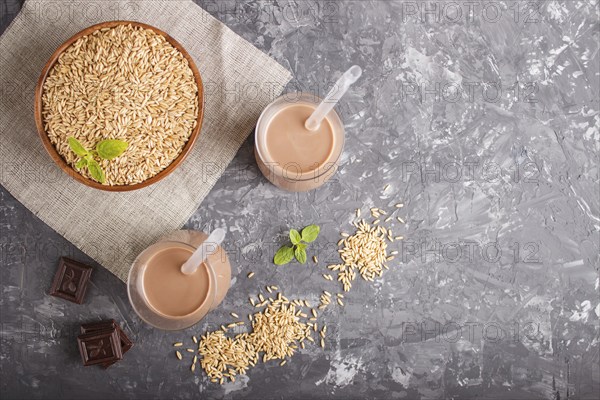 Organic non dairy oat chocolate milk in glass and wooden plate with oat seeds on a gray concrete background. Vegan healthy food concept, flat lay, top view, copy space