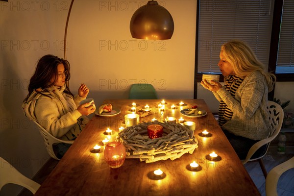 Symbolic image energy saving, cold winter, energy crisis, cold flat, mother and daughter dressed warmly at dinner, only by candlelight, heating on to a minimum