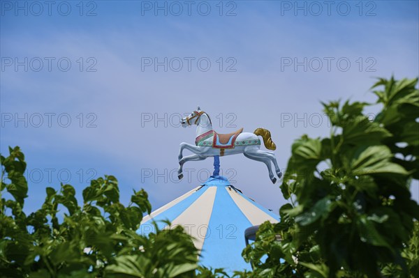 A carousel horse stands on a blue and white striped roof, surrounded by green leaves under a clear sky, Saintes-Maries-de-la-Mer, Camargue, France, Europe