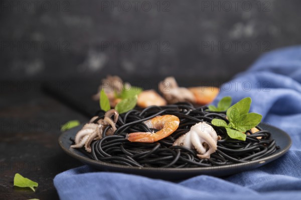 Black cuttlefish ink pasta with shrimps or prawns and small octopuses on black concrete background and blue textile. Side view, close up, selective focus