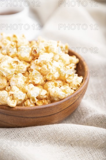 Popcorn with caramel in wooden bowl and a cup of coffee on a white wooden background and linen textile. Side view, close up, selective focus