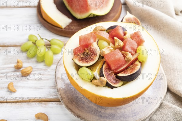 Vegetarian fruit salad of watermelon, grapes, figs, pear, orange, cashew on white wooden background and linen textile. Side view, close up