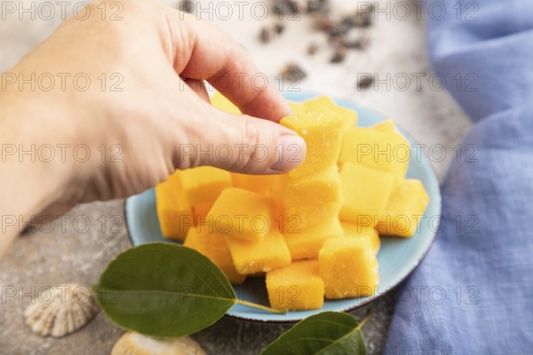 Dried and candied mango cubes on blue plate with hand on brown concrete background and linen textile. Side view, close up, selective focus, vegan, vegetarian food concept