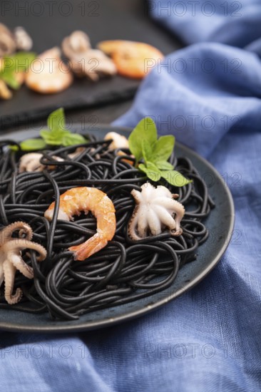 Black cuttlefish ink pasta with shrimps or prawns and small octopuses on black concrete background and blue textile. Side view, close up, selective focus