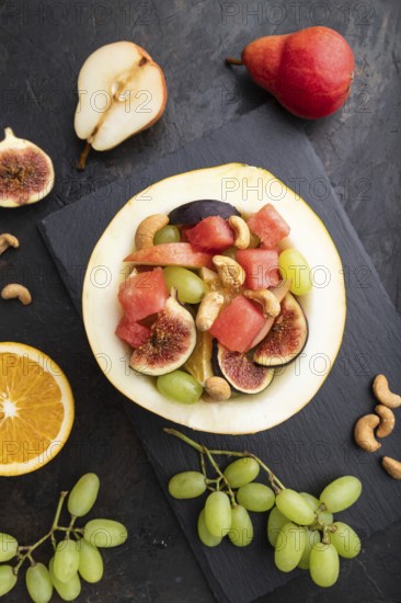 Vegetarian fruit salad of watermelon, grapes, figs, pear, orange, cashew on slate board on a black concrete background. Top view, flat lay, close up