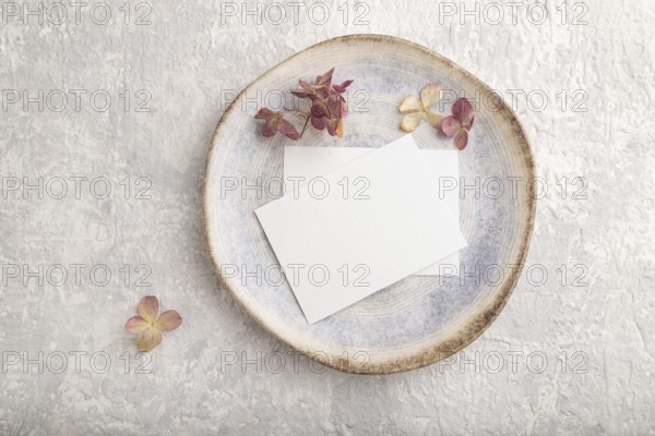 White paper invitation card, mockup with dried hydrangea flowers on ceramic plate and gray concrete background. Blank, flat lay, top view, still life, copy space