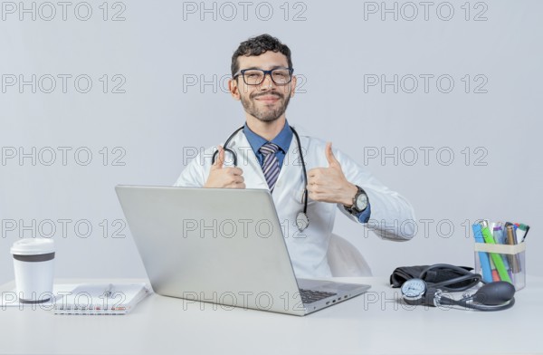 Smiling doctor sitting with laptop giving thumbs up. Male doctor with laptop showing thumbs up in ok gesture