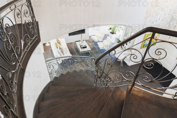 Looking down spiraling wooden staircase with wrought iron balusters onto entry way and living room with white leather sofa and armchairs, glass-top coffee table inside a luxurious residential home, Quebec, Canada, North America