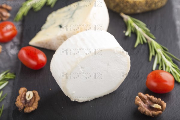 White goat cheese and various types of cheese with rosemary and tomatoes on black slate board on a black concrete background. Side view, close up, selective focus