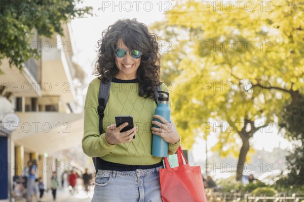 Smiling woman with curly hair walks through the city at sunset, holding a water bottle and red bag. Dressed in a green sweater and jeans, she enjoys a relaxed stroll in the warm evening light