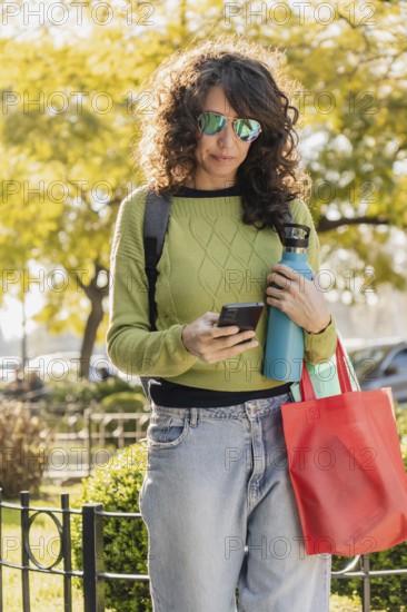 Woman with curly hair and sunglasses uses her smartphone in a park at sunset. Dressed casually in a green sweater and jeans, she holds a water bottle and a red bag, enjoying the outdoor environment