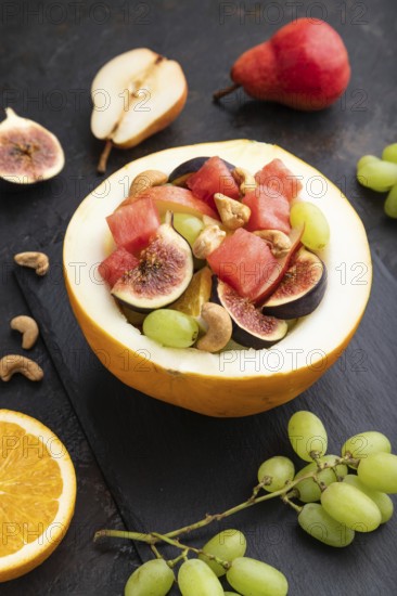 Vegetarian fruit salad of watermelon, grapes, figs, pear, orange, cashew on slate board on a black concrete background. Side view, close up