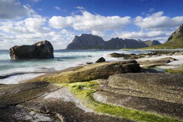 Seascape on the beach at Uttakleiv (Utakleiv), rocks and green seaweed in the foreground. In the background the mountain Hogskolmen. Good weather, blue sky with some clouds. Early summer. Long exposure. Uttakleiv, Vestvagoya, Lofoten, Norway, Europe