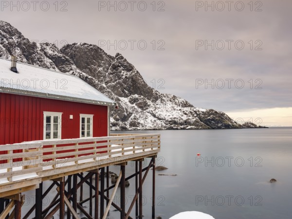 Traditional red wooden house on pegs by the fjord, snow-capped mountains behind, Nusfjord, Flakstadøya, Lofoten, Norway, Europe