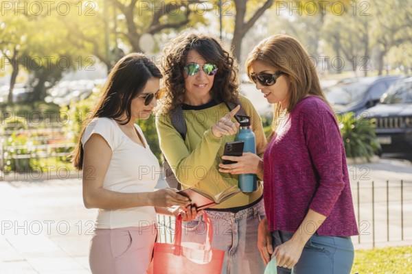 Three women standing outdoors, smiling and interacting with a smartphone. The group enjoys a sunny day, chatting and interacting with technology while wearing casual, colorful clothing and wearing sunglasses