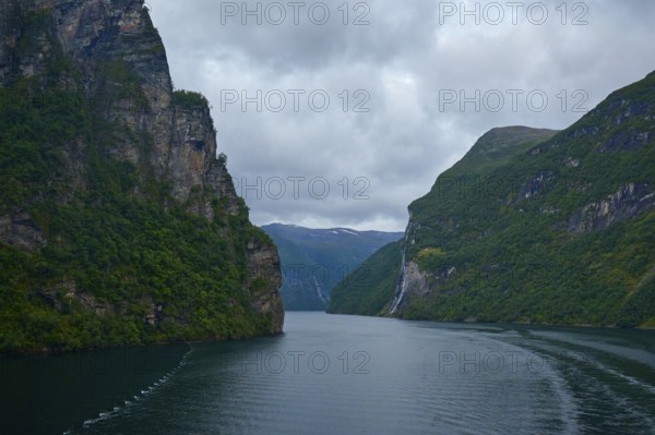 A fjord surrounded by rocky mountains under a cloudy sky with a waterfall in the background, Geiranger, Geiranger Fjord, Stranda, Romsdal, Norway, Europe