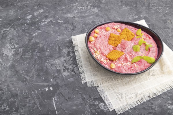 Hummus with beet and microgreen basil sprouts in ceramic bowl on a black concrete background and linen textile. Side view, copy space