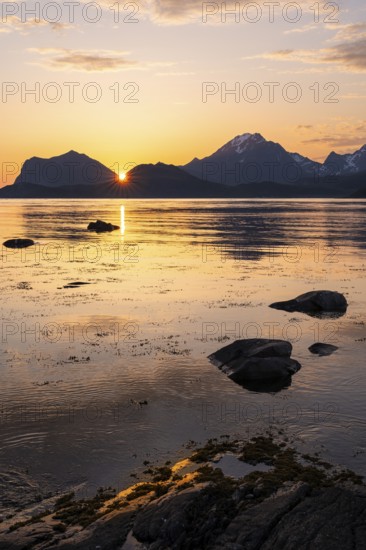 Landscape on the Lofoten Islands. View from Flakstadoya to Vestvagoya, with the snow-covered Himmeltindan mountain on the right. The mountains are reflected in the sea. The sun is between two mountains. At night at the time of the midnight sun in good weather. Early summer. Lofoten, Norway, Europe