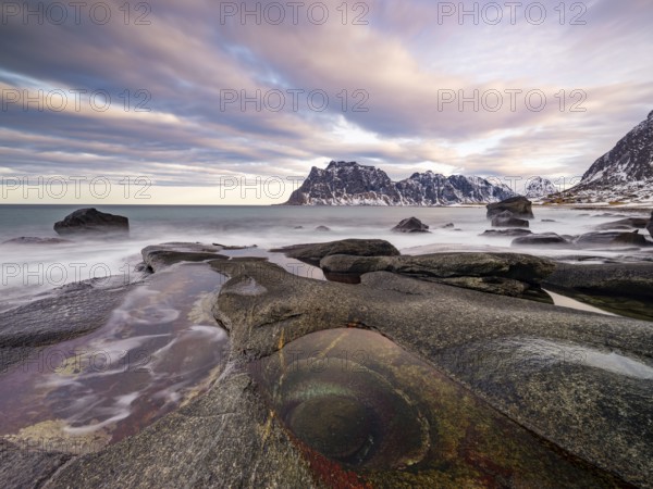 Rocks at Utakleiv beach with rock formation Eye of Utakleiv, or Dragon's Eye, in a dramatic cloudy atmosphere, snow-covered mountains in the background, Vestvågøya, Lofoten, Norway, Europe