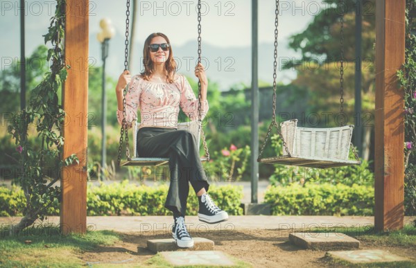 Cheerful girl in glasses relaxing on a swing in a natural park. Happy young woman sitting on a swing in a nature park