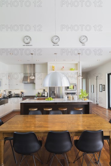 Open-concept dining and kitchen area with antique dining table with black leather and plastic chairs, marble top wooden island with painted black steel and exotic wood bar stools, white cabinets inside a luxurious modern cube style house, Quebec, Canada, North America