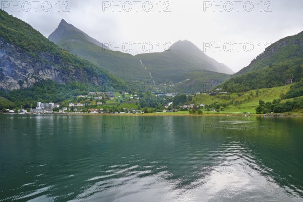 Mountainous landscape in a fjord reflecting the surrounding mountains and village, surrounded by green nature and under a cloudy sky, Geiranger, Geiranger Fjord, Stranda, Romsdal, Norway, Europe