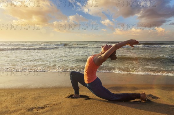 Yoga outdoors, sporty fit woman practices yoga Anjaneyasana, low crescent lunge pose outdoors at beach on sunset