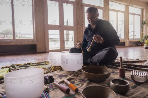 A mature Caucasian man performing with a quartz singing bowl amidst a collection of vibrational instruments for sound meditation sessions